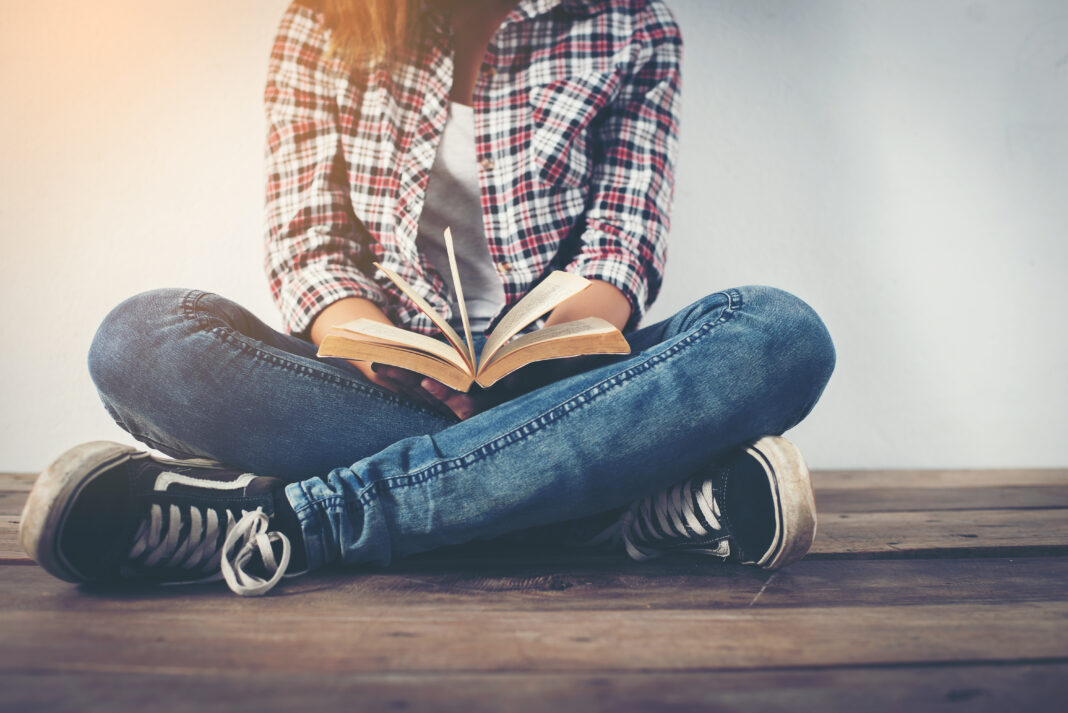 Young hipster woman holding open book sitting on wooden floor.